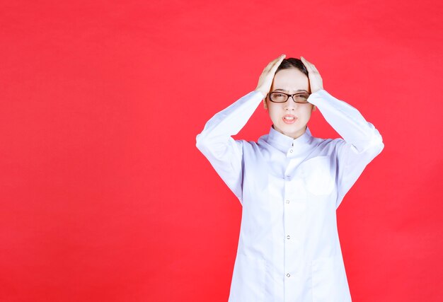 Female doctor in eyeglasses standing on red background holding head with hands and looks terrified.