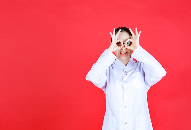 Free photo female doctor in eyeglasses standing on red background and looking across her fingers.