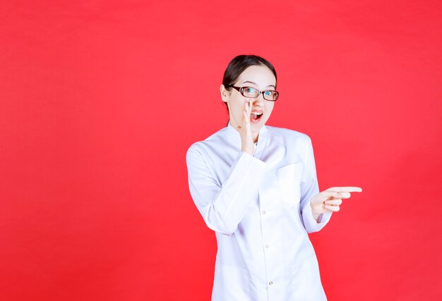 Female doctor in eyeglasses standing on red background and showing right side.