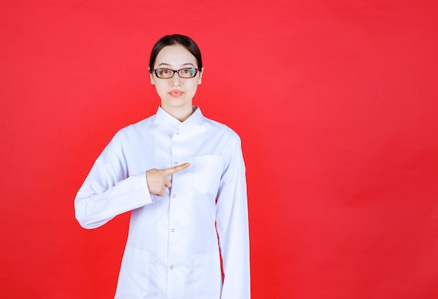 Female doctor in eyeglasses standing on red background and showing the right side.