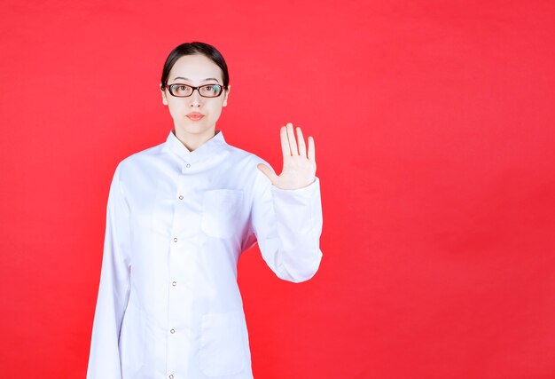 안경을 쓴 여성 의사는 빨간색 배경에 서서 손으로 무언가를 멈추고 있습니다.