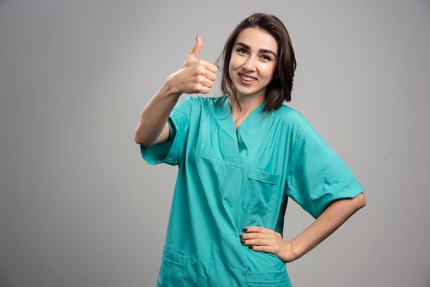 Женщина-врач дает большие пальцы руки вверх на сером фоне. Фото высокого качества