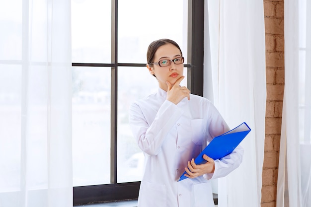 Female doctor in glasses holding blue folder near window. 