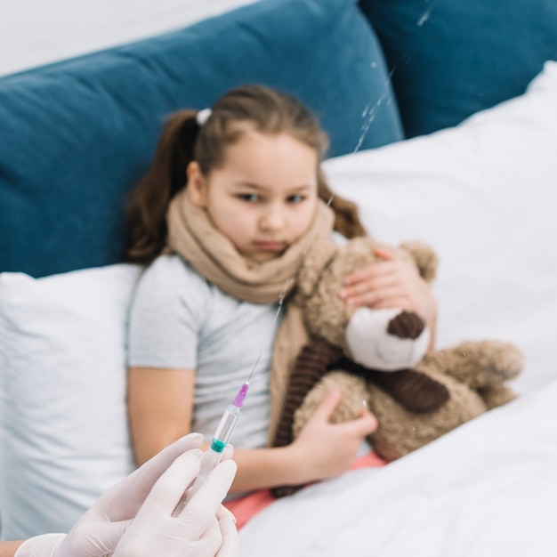 Female doctor hands spraying the syringe with medicine in front of sick girl sitting with teddy bear on bed