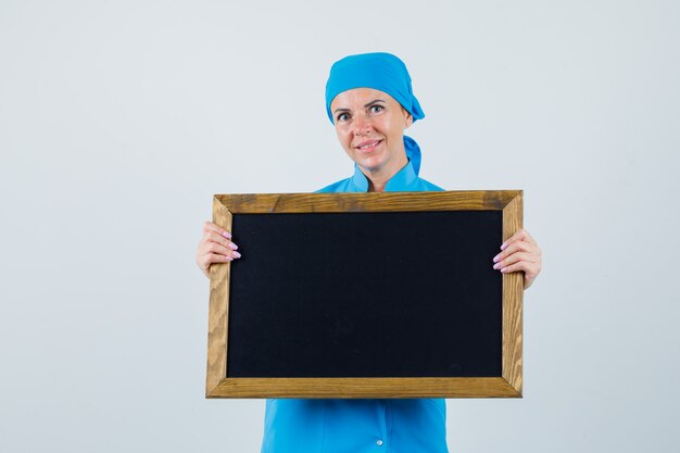 Female doctor holding blackboard in blue uniform and looking optimistic , front view.