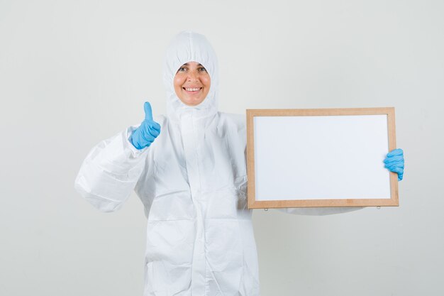 Female doctor holding blank frame with thumb up in protection suit