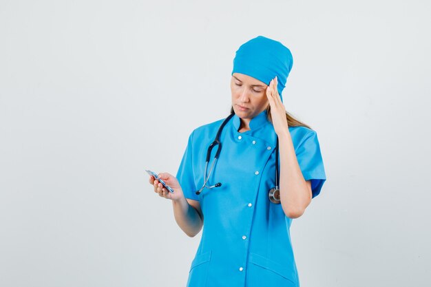 Female doctor holding capsules with fingers on temples in blue uniform and looking painful. front view.