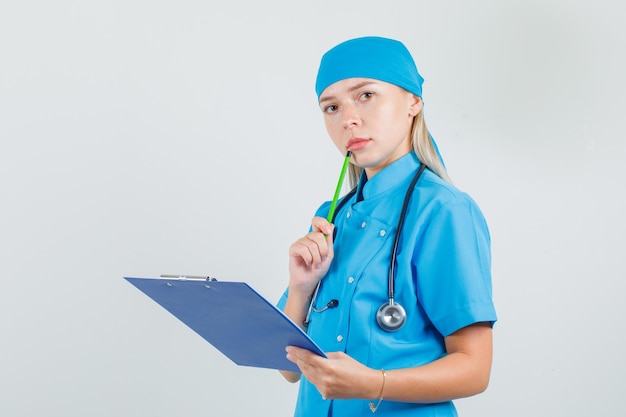 青い制服を着た口の近くに鉛筆でクリップボードを保持し、忙しそうに見える女性医師