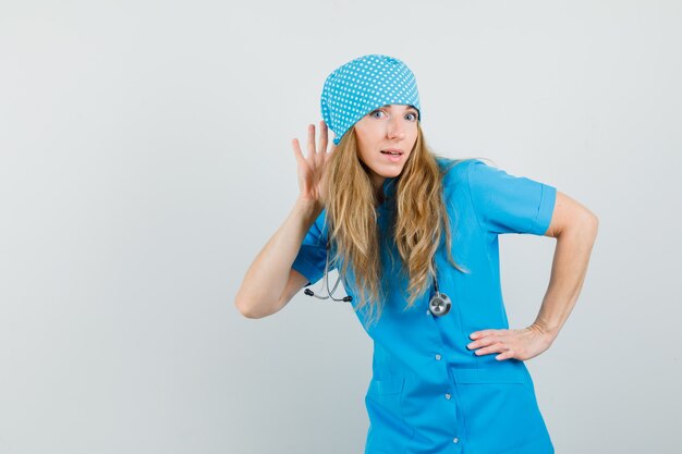 Female doctor holding hand behind ear to listen in blue uniform and looking curious 
