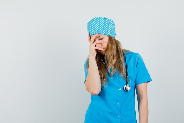Женщина-врач держит руку на лице в синей форме и выглядит усталой.