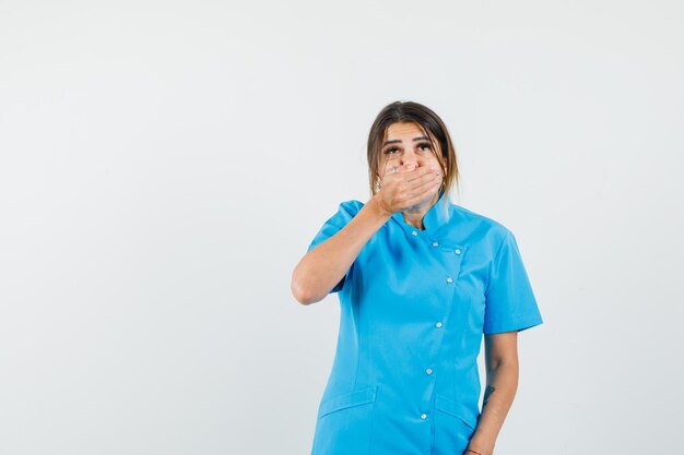 Female doctor holding hand on mouth in blue uniform and looking scared