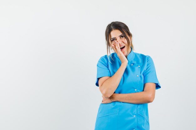 Female doctor holding hand near open mouth in blue uniform and looking amazed