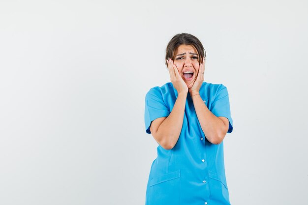 Female doctor holding hands on cheeks in blue uniform and looking wistful