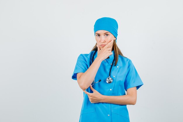 파란색 유니폼을 입고 생각하고 심각한 찾고있는 동안 그녀의 턱을 잡고 여성 의사. 전면보기.
