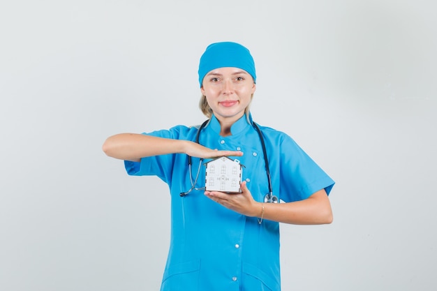 Женщина-врач держит модель дома и улыбается в синей форме