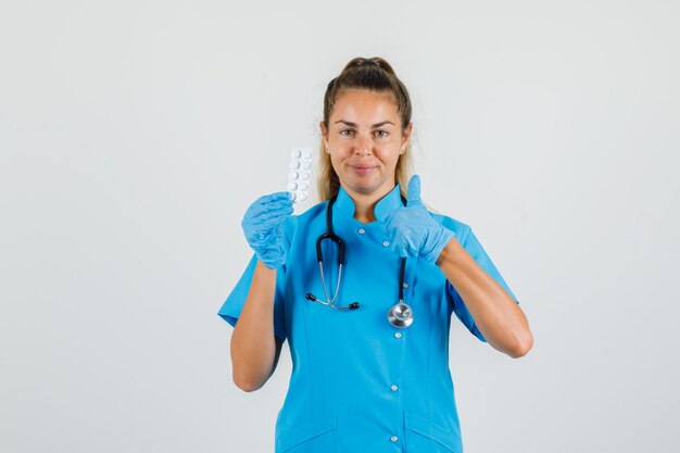 Female doctor holding pack of pills with thumb up in blue uniform, gloves and looking positive