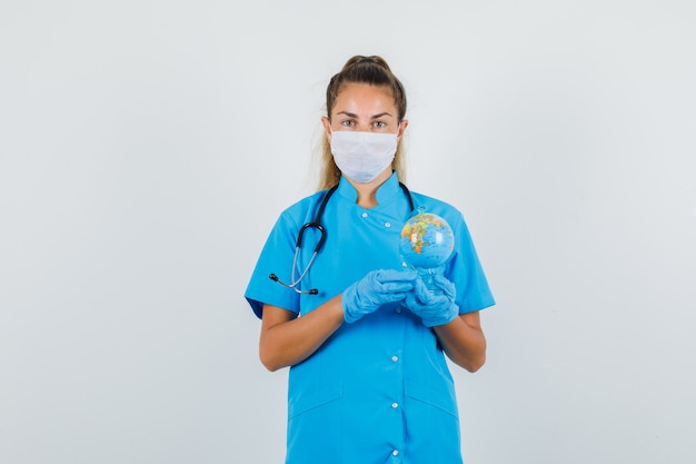Medico femminile che tiene il globo del mondo in uniforme blu, maschera, guanti e guardando attento.