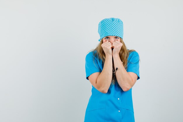 Female doctor keeping fists on face in blue uniform and looking scared. 