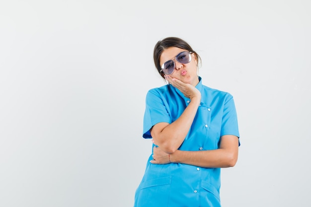 여성 의사가 입술을 접고, 파란색 유니폼을 입은 손바닥에 뺨을 기대고 있습니다.