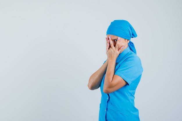 Female doctor looking through fingers in blue uniform and looking curious , front view.