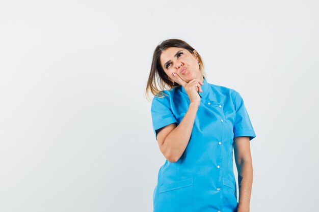 Женщина-врач смотрит вверх, надувает губы в синей форме и выглядит задумчиво