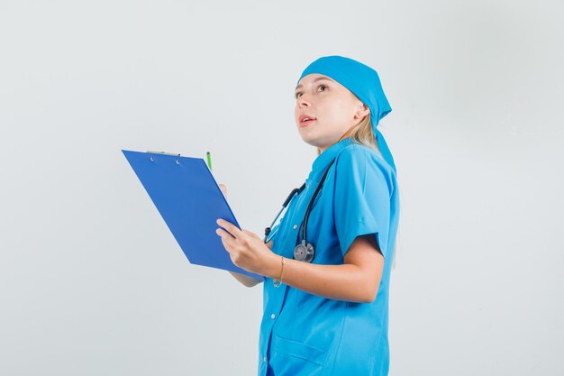 Female doctor looking up while holding clipboard and pencil in blue uniform