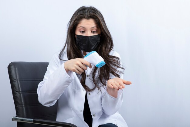 Female doctor in medical mask checking temperature on white wall.