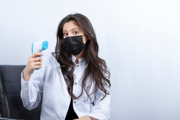 Female doctor in medical mask holding thermometer and looking at front. 