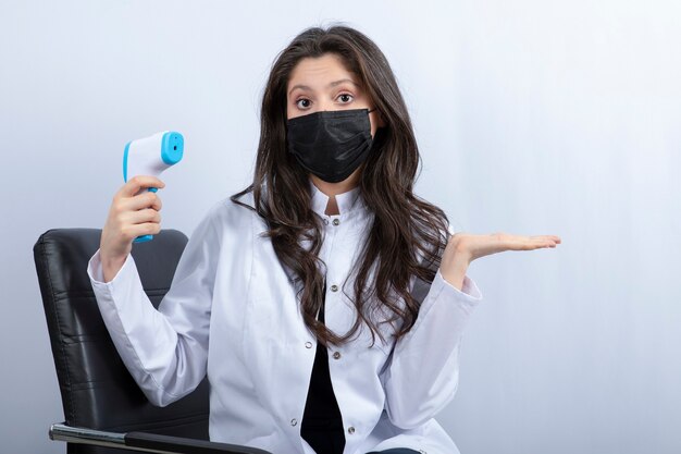 Female doctor in medical mask holding thermometer and looking at front. 