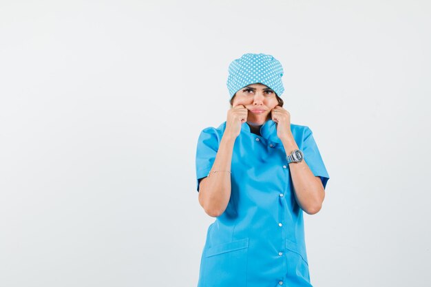 Женщина-врач щипает щеки в синей форме и выглядит мрачно