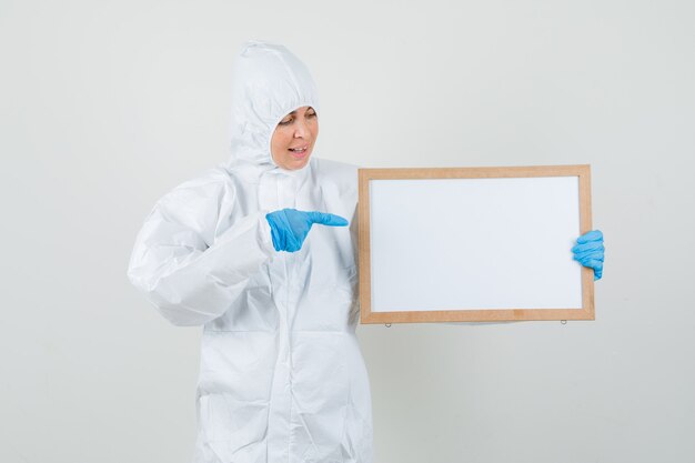 Женщина-врач указывая на пустую рамку в защитном костюме, перчатках и весело глядя.