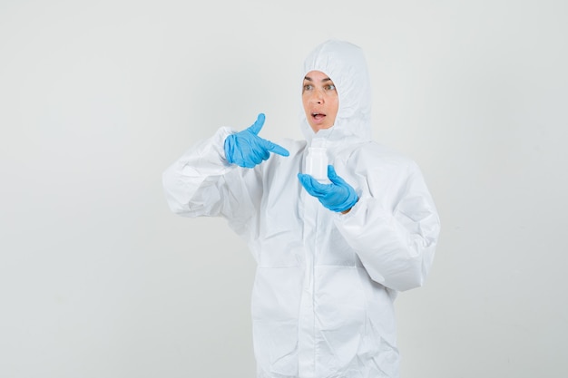 Female doctor pointing at bottle of pills in protective suit, gloves and looking confused