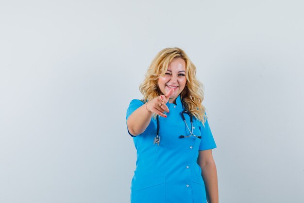 Female doctor pointing at camera while winking in blue uniform and looking amused.