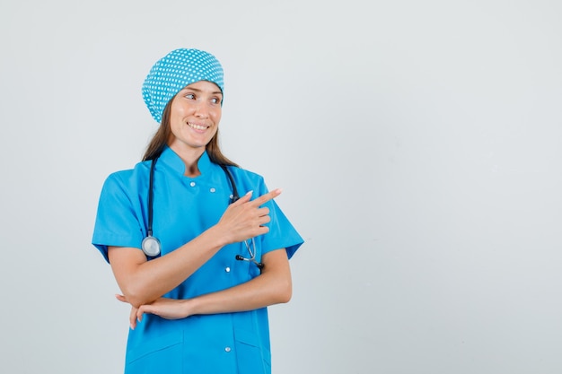 Female doctor pointing finger away in blue uniform and looking cheerful. front view