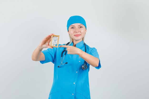 Женщина-врач указывая пальцем на песочные часы в синей форме и выглядит веселым.