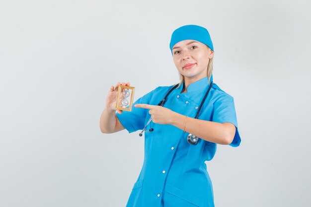 Female doctor pointing finger at hourglass in blue uniform and looking cheerful.