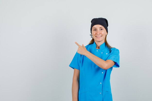 Female doctor pointing finger to side in blue uniform, black hat and looking cheerful