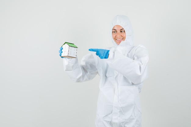 Female doctor pointing at house model in protective suit, gloves and looking optimistic.