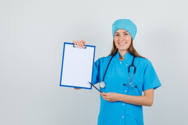 Female doctor pointing pen at clipboard in blue uniform and looking glad. front view.