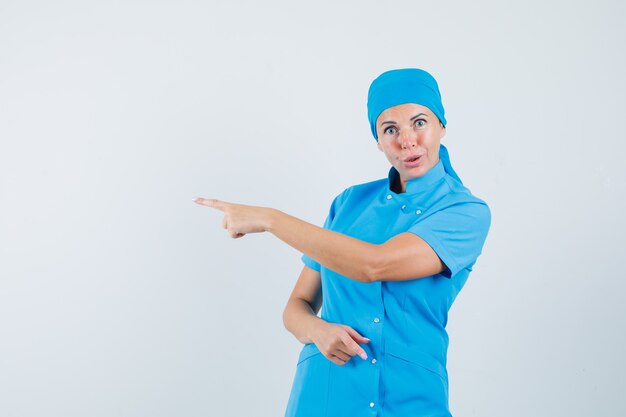 Female doctor pointing to the side in blue uniform and looking hesitant. front view.