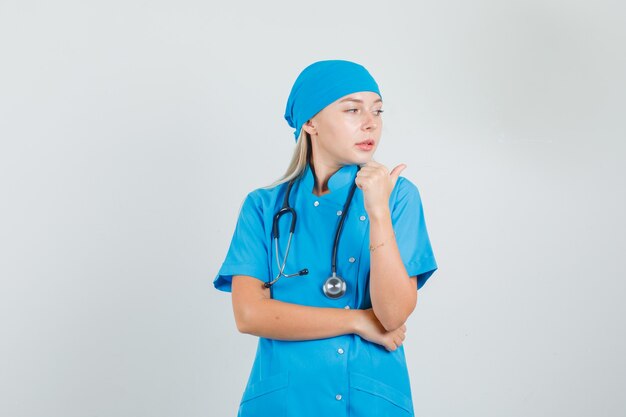 Female doctor pointing to side with thumb in blue uniform and looking cute