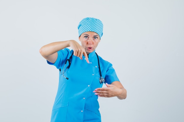 Female doctor pointing at something pretended to be held in blue uniform and looking surprised , front view