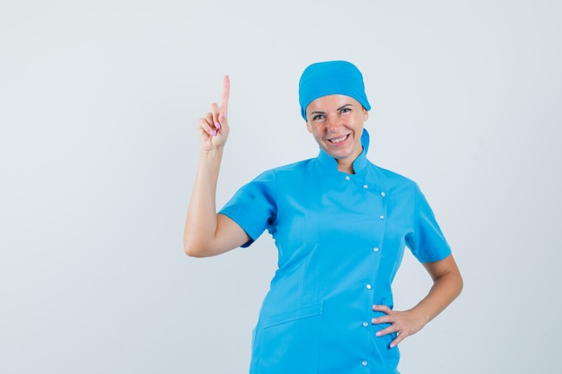 Женщина-врач, указывая вверх в синей форме и глядя веселый, вид спереди.