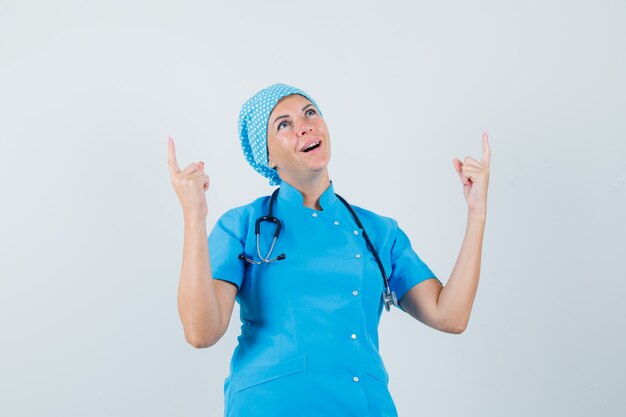 Female doctor pointing up in blue uniform and looking grateful , front view.