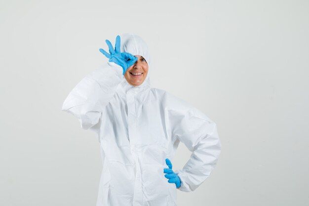 Female doctor in protective suit, gloves showing ok sign on eye and looking funny