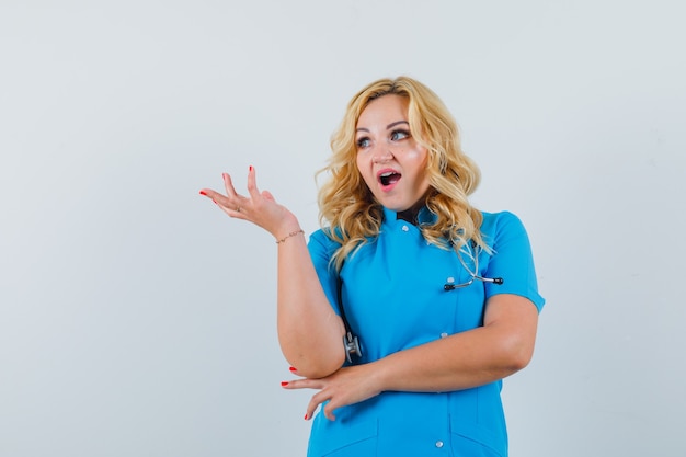 Женщина-врач поднимает руку во время разговора в синей форме и смотрит сосредоточенным