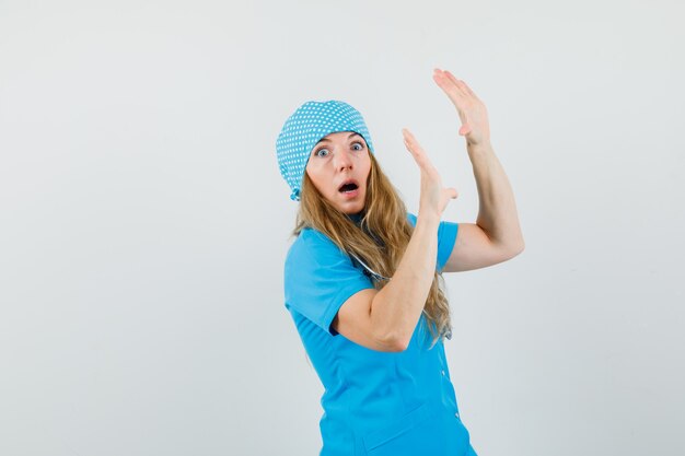 Female doctor raising palms to defend herself in blue uniform and looking scared 