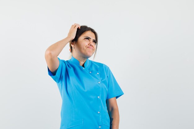 파란색 유니폼을 입고 우유부단하게 보는 동안 여성 의사가 머리를 긁적