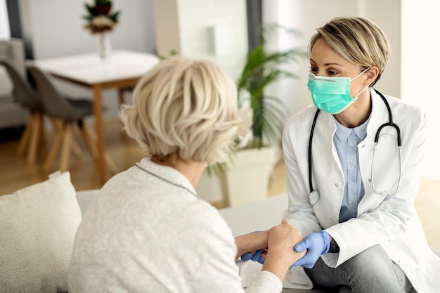 Female doctor and senior woman wearing face masks while holding hands and communicating during home visit