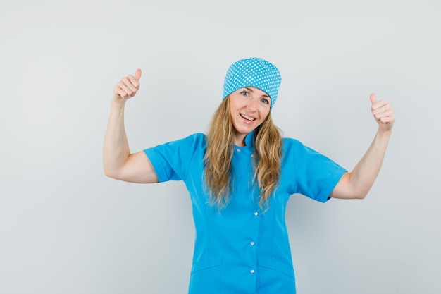 Женщина-врач показывает двойные пальцы вверх в синей форме и выглядит счастливой.